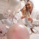Zwei hübsche junge Frauen feiern ihren JGA bei Regen mit Sekt auf einem Bett mit weißem Laken im Vordergrund sind pinke Luftballons zu sehen